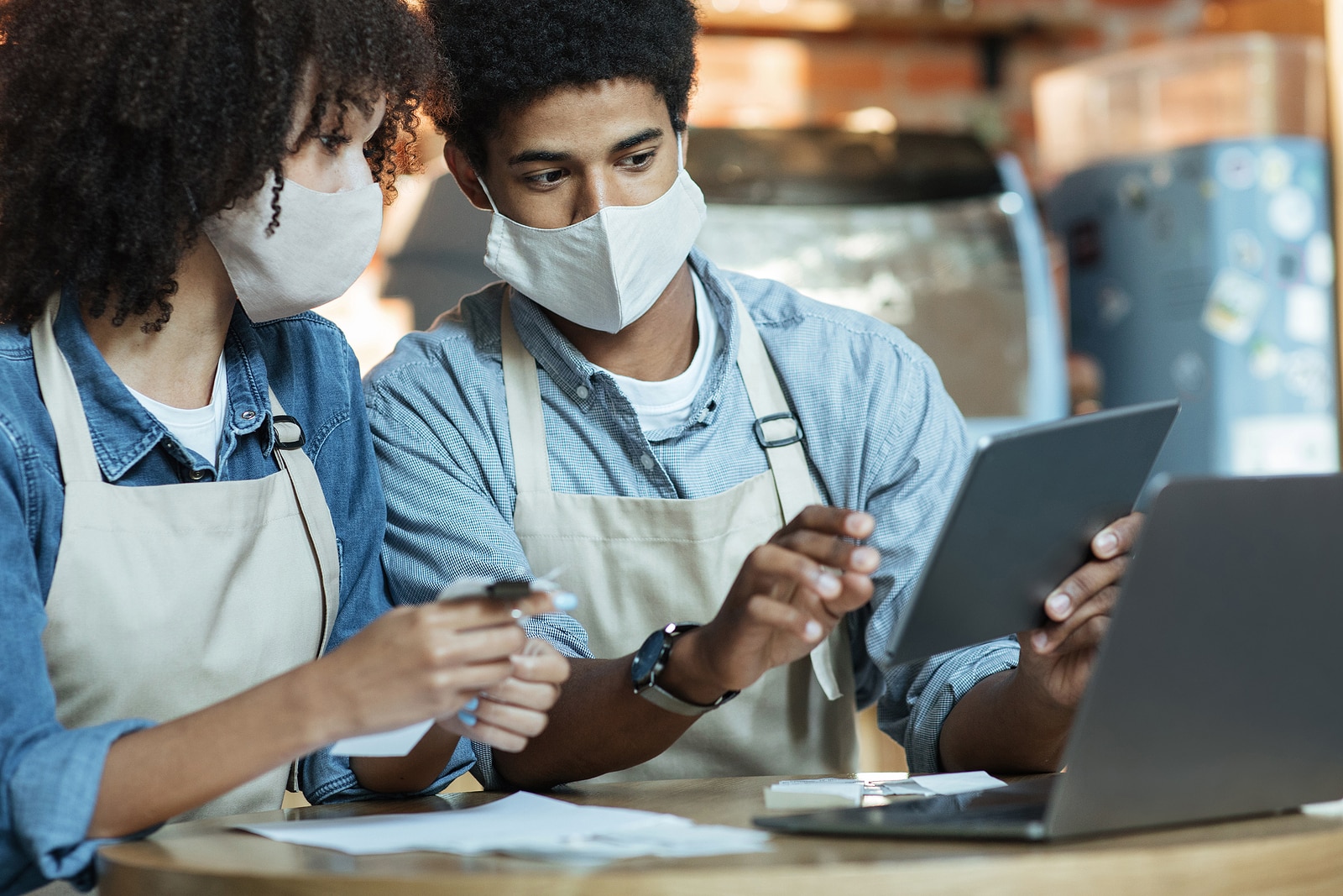 Small business owners at social distancing of covid quarantine. Busy african american couple in aprons and protective masks work with laptop and tablet to attract clients in trendy cafe or restaurant